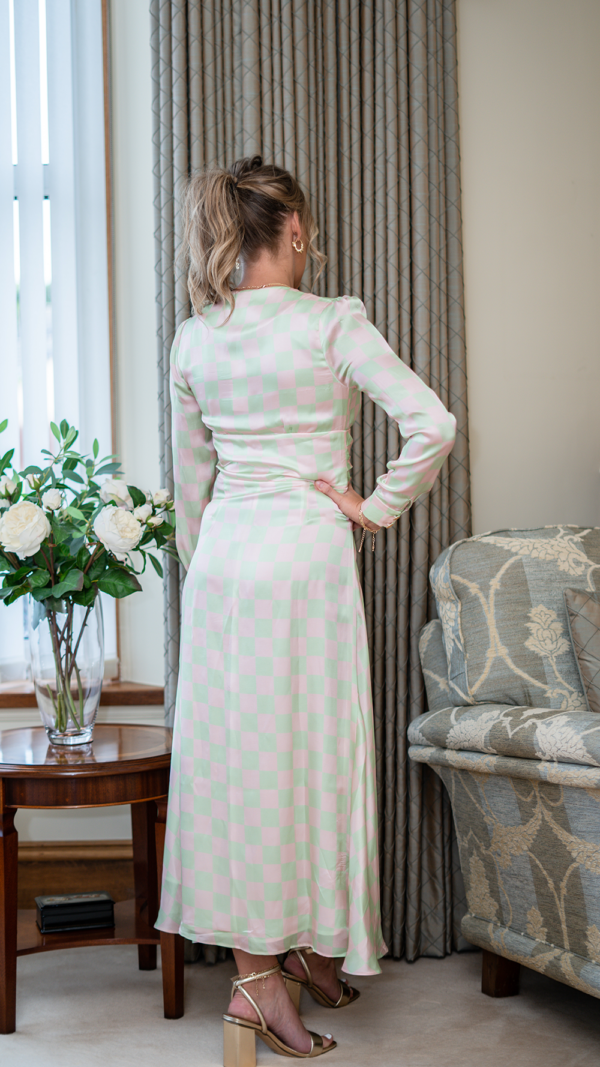 Model is tall with wavy blonde hair in a ponytail. Her back is facing the camera and she wears the Olivia Rubin Lliana dress, which has a pale pink and green checked patter. It is a long dress, falling almost to the ankles, and has long sleeves.