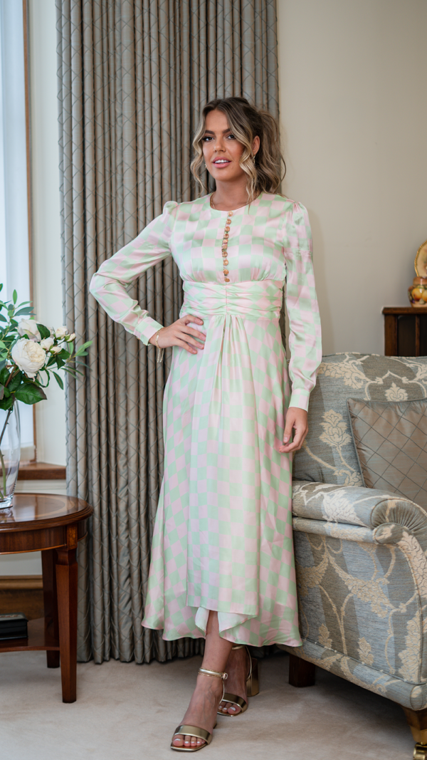 Model is tall with wavy blonde hair in a ponytail. She is facing the camera, with one hand resting on her hip. She she wears the Olivia Rubin Lliana dress, which has a pale pink and green checked patter. It is a long dress, falling almost to the ankles, has buttons trailing down from the neckline and has long sleeves.