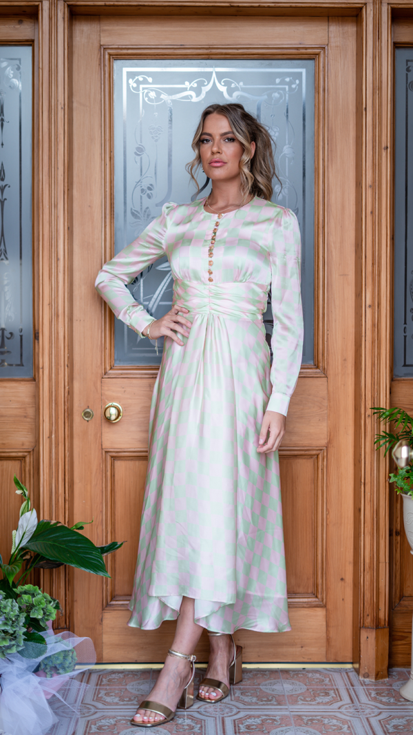 Model is tall with wavy blonde hair in a ponytail. She is facing the camera, with one hand resting on her hip. She she wears the Olivia Rubin Lliana dress, which has a pale pink and green checked patter. It is a long dress, falling almost to the ankles, has buttons trailing down from the neckline and has long sleeves.
