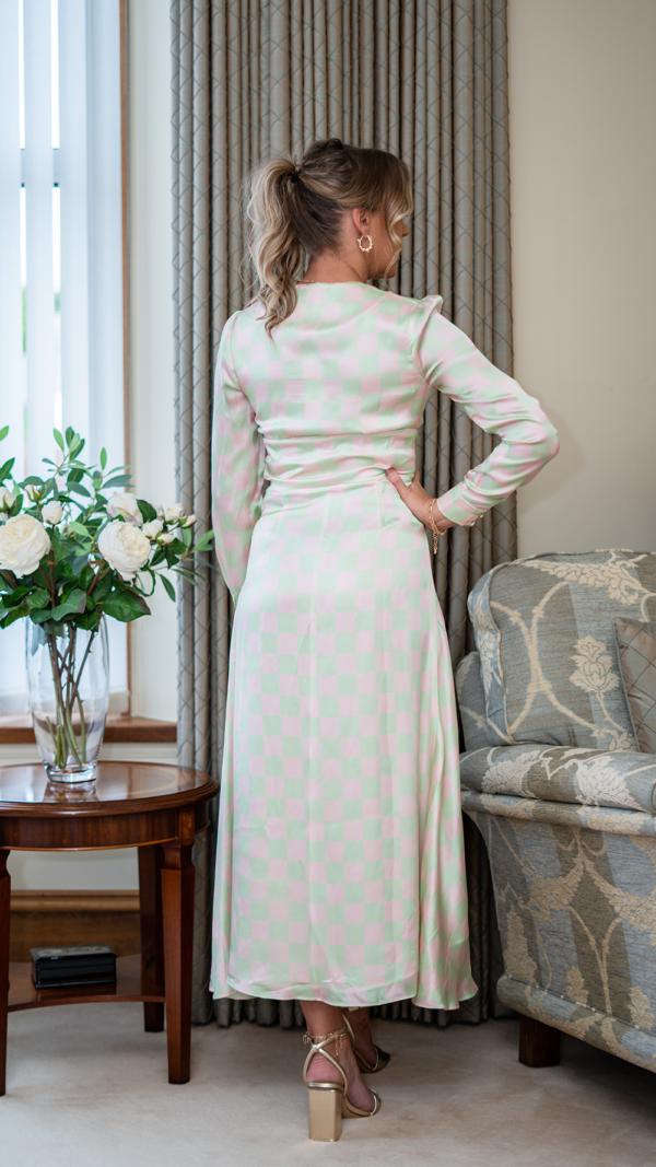 Model is tall with wavy blonde hair in a ponytail. Her back is facing the camera and she wears the Olivia Rubin Lliana dress, which has a pale pink and green checked patter. It is a long dress, falling almost to the ankles, and has long sleeves.