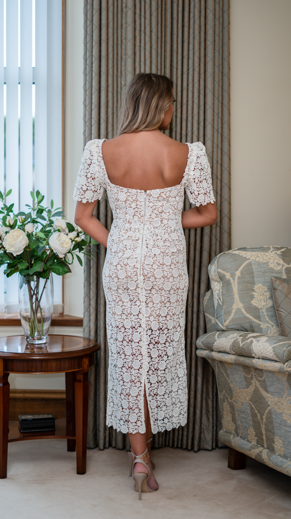 Model is tall with long wavy blonde hair. Her back faces the camera and she is wearing the white lace Guipure dress. It is long, with short sleeves.