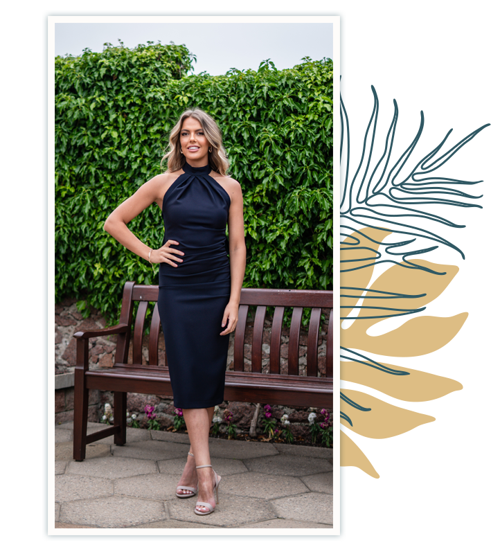 Model is tall with wavy blonde hair tied in a ponytail. She stands outside, in front of a wooden bench and lush green foliage. She wears the Kevan Jon Akash dress in navy blue. It is a midi dress, with wide straps crossing at the neckline and reaching around the upper arms.