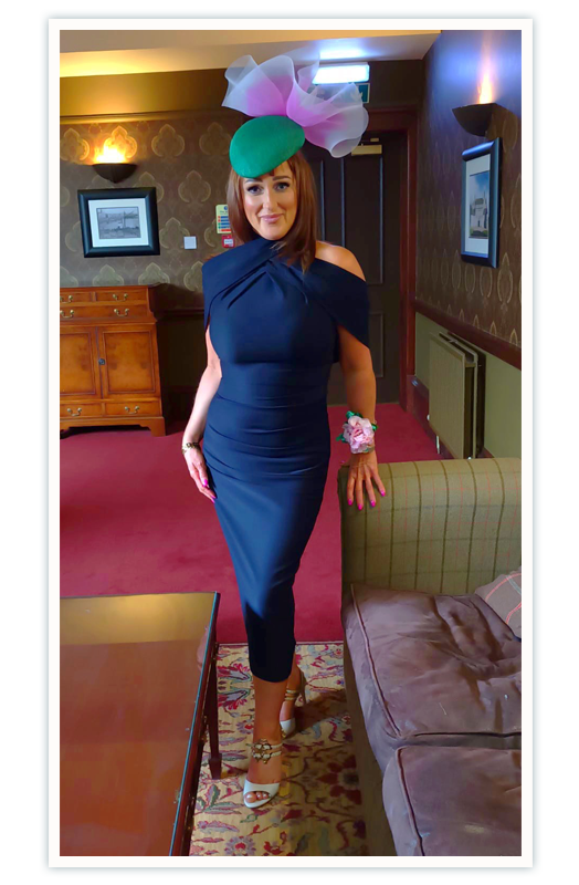 Fiona, the founder of Island Palm Designer Dress Hire, is tall and slender with dark, shoulder length hair. She smiles at the camera and is wearing a navy blue dress, a green hat with pink frill decoration, and a flower corsage on her wrist.